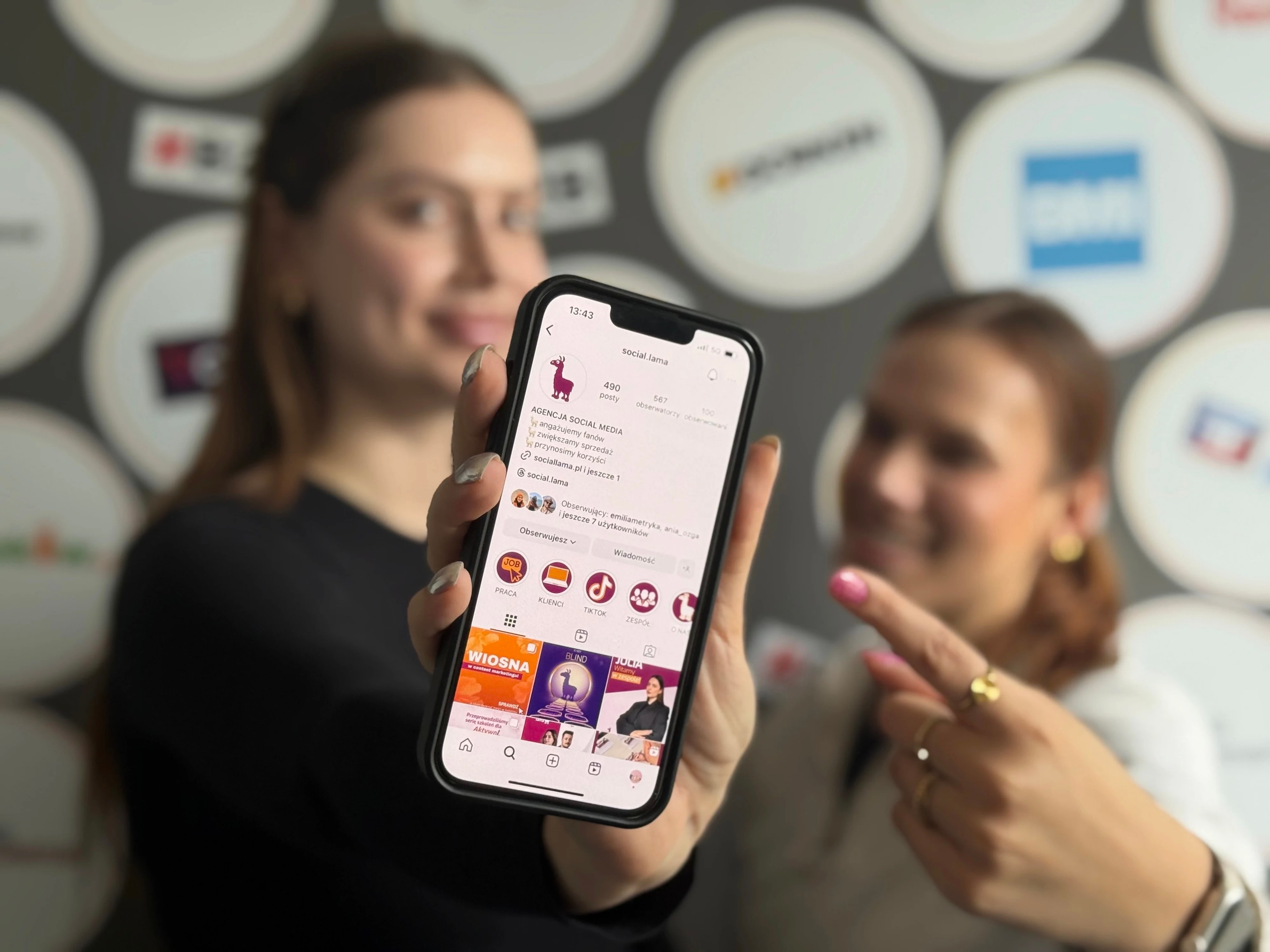 Two Social Lama staff holding up a phone showing the @social.lama Instagram profile, its bio reading "AGENCJA SOCIAL MEDIA — angażujemy fanów, zwiększamy sprzedaż, przynosimy korzyści" ("social media agency — we engage fans, grow sales, deliver results"), against a wall of client logos.